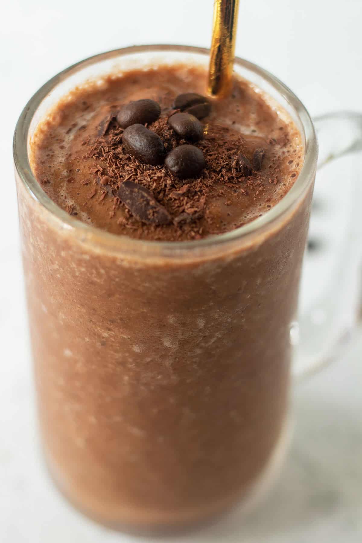 mocha coffee smoothie recipe in a clear glass garnished with shaved chocolate, fresh coffee beans and a gold straw for serving.