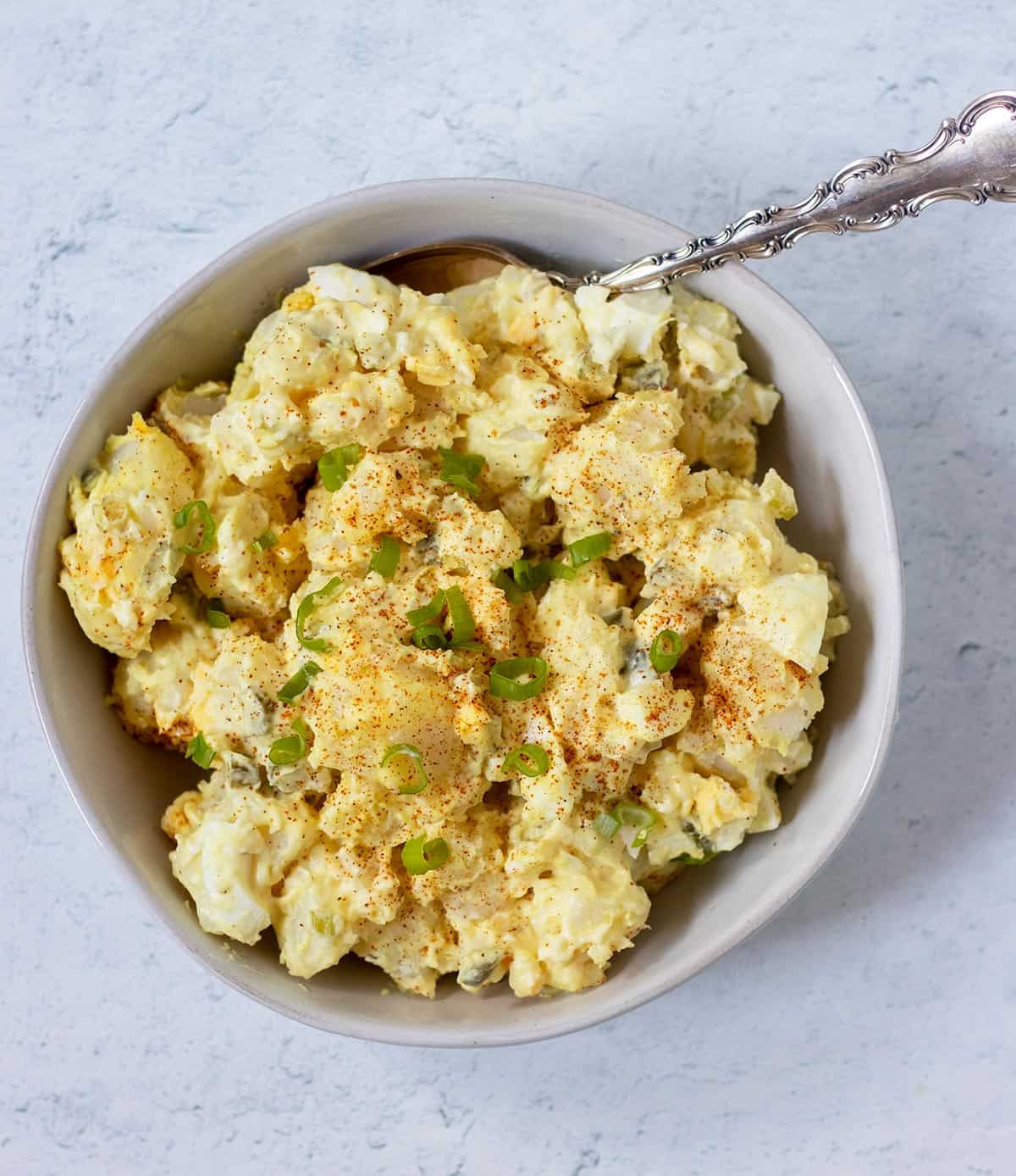mustard potato salad in a white bowl with a silver spoon that's garnished with paprika and chopped green onions.