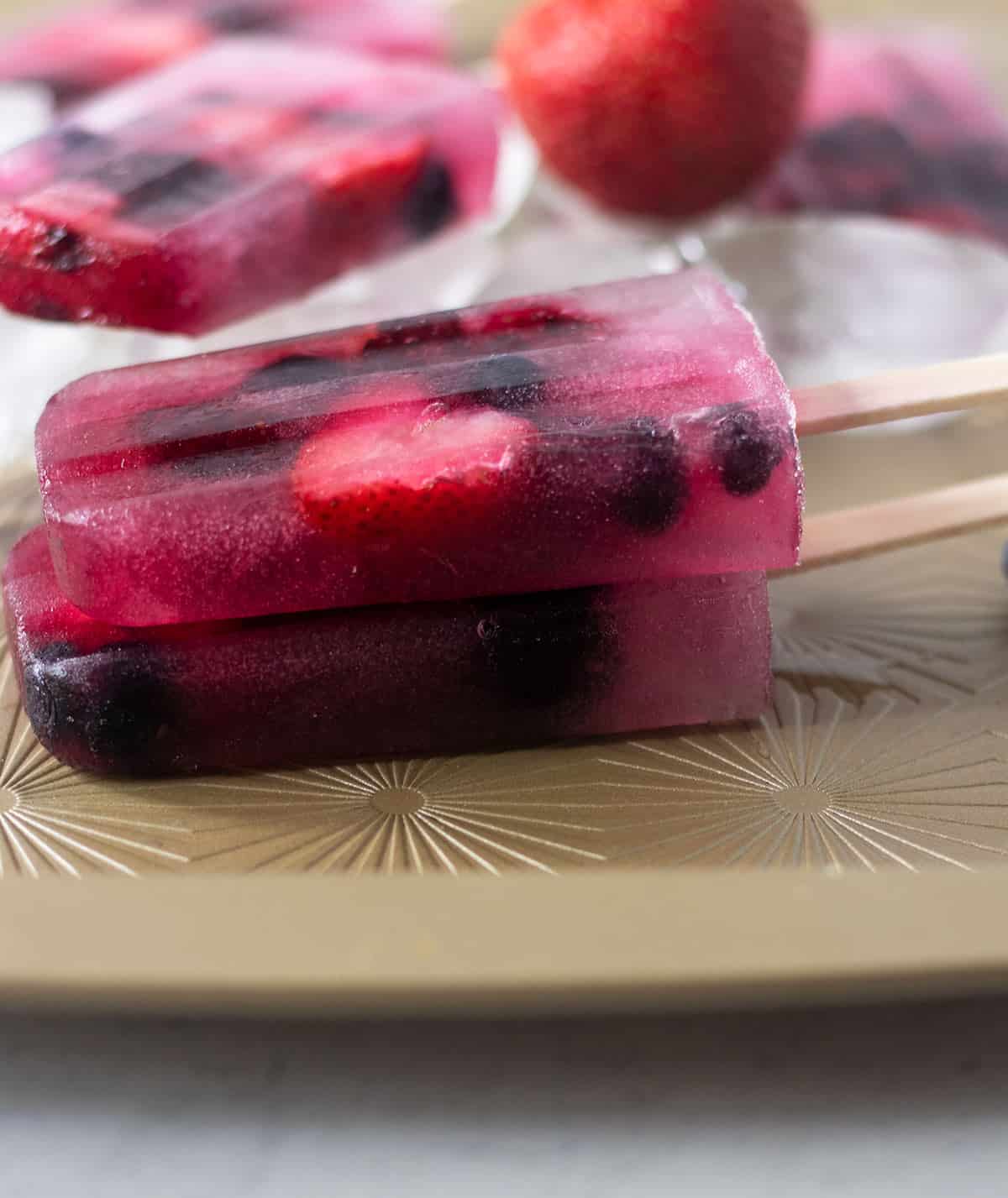 two mixed fruit popsicles stacked on top of each other on a gold tray with more popsicles and ice in the background. Also a fresh strawberry in the background.