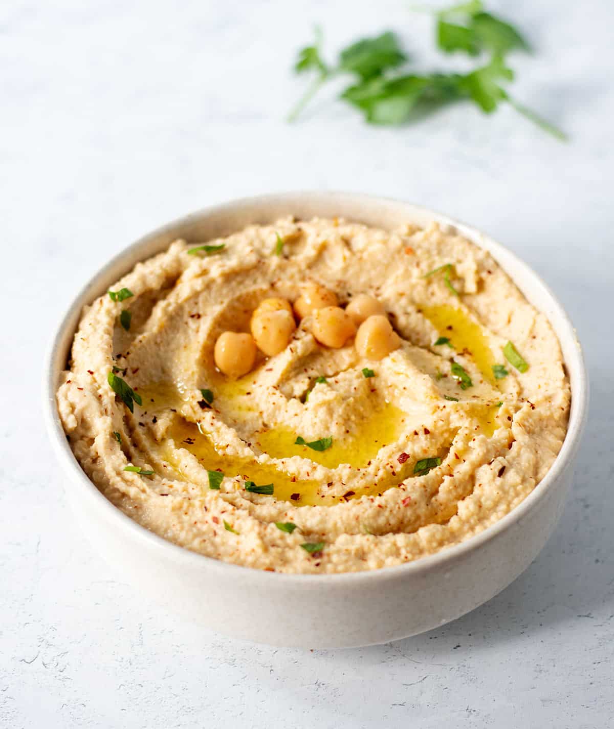 roasted garlic hummus in a white bowl garnished with fresh chopped parsley, chickpeas, olive oil and red chili pepper flakes.