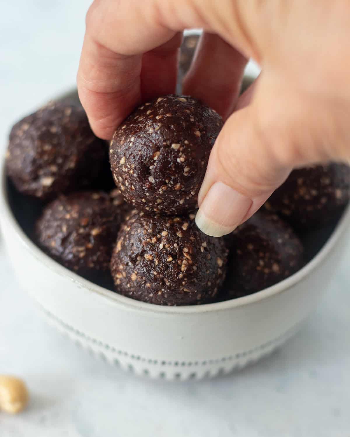 chocolate protein balls in a white bowl with blue trim and a hand reaching in to pick one up. A cashew it beside the bowl.
