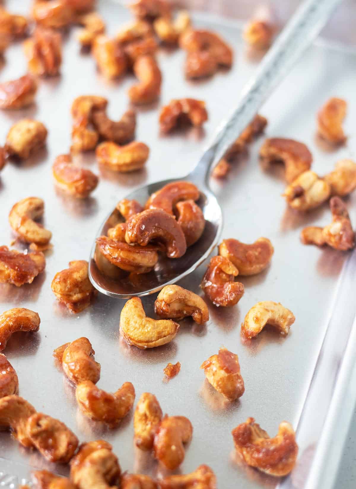 honey roasted cashews on a baking pan after being roasted with a silver spoon scooping some nuts up.