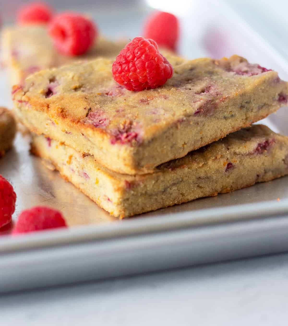 two raspberry scones stacked on a baking sheet with fresh raspberries on top.