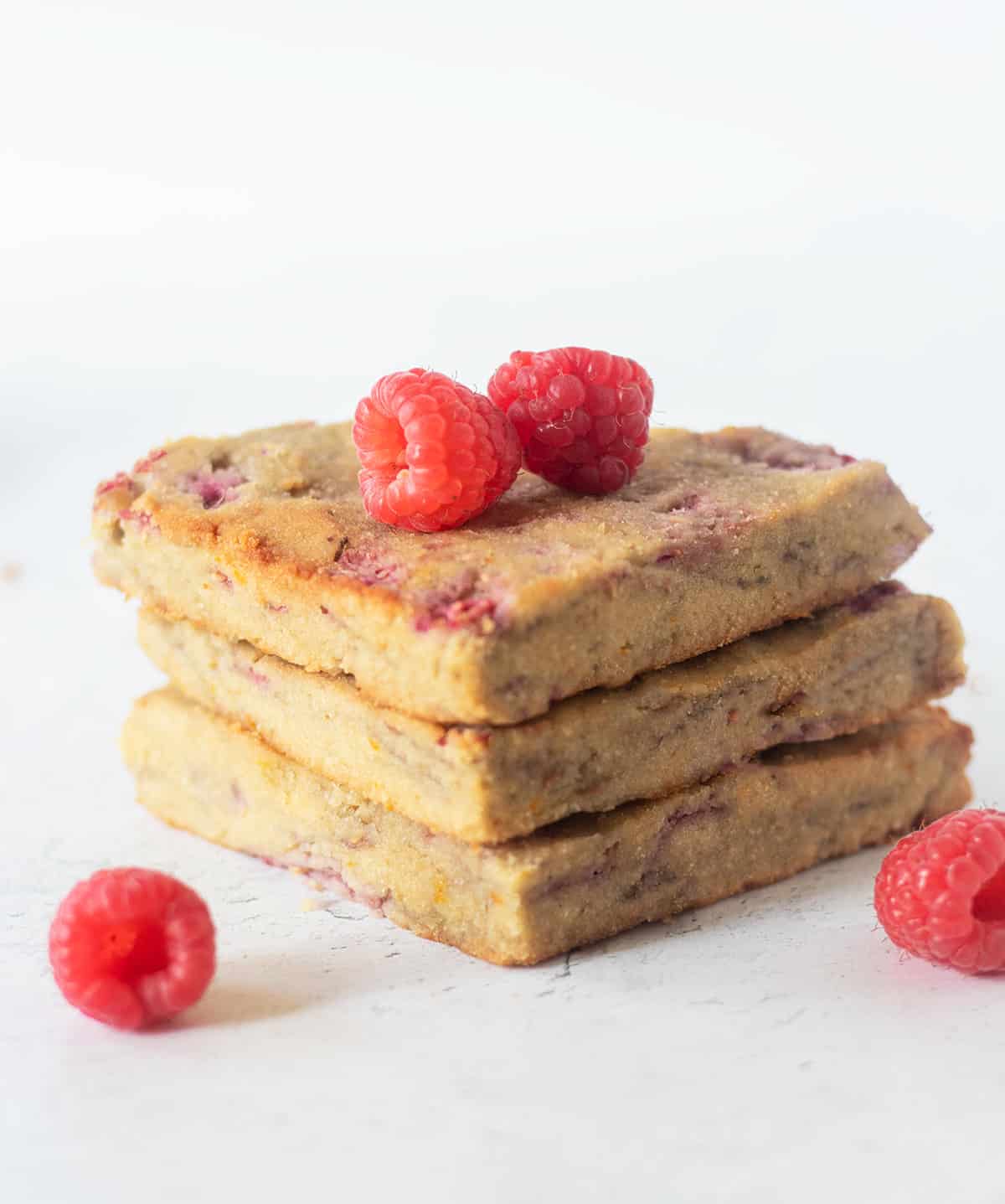 three raspberry scones stacked on top of each other with fresh raspberries beside them and on top of them.