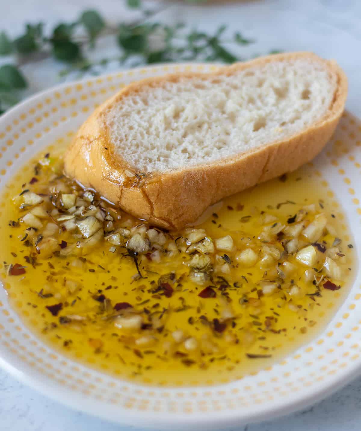 garlic bread dipping oil on a white and yellow plate with a slice of bread and fresh oregano in the background.
