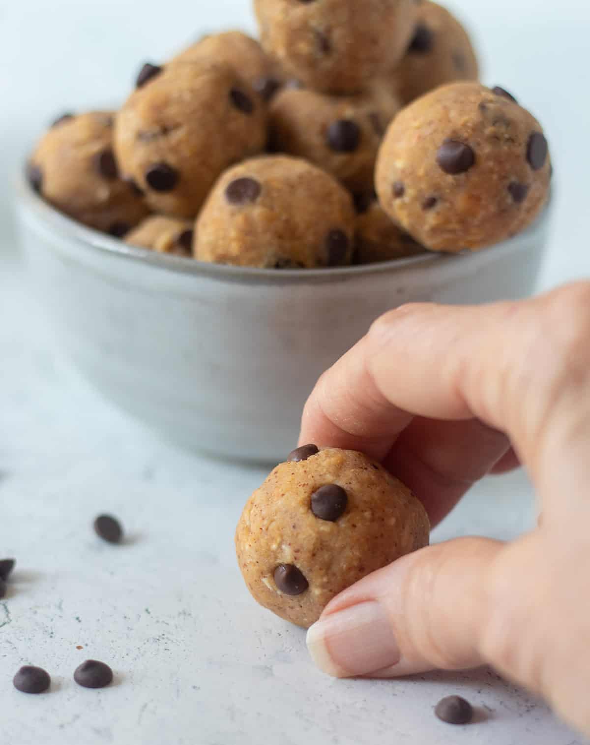 A hand grabbing a chickpea cookie dough bite with a bowl of them in the background and mini chocolate chips beside them.