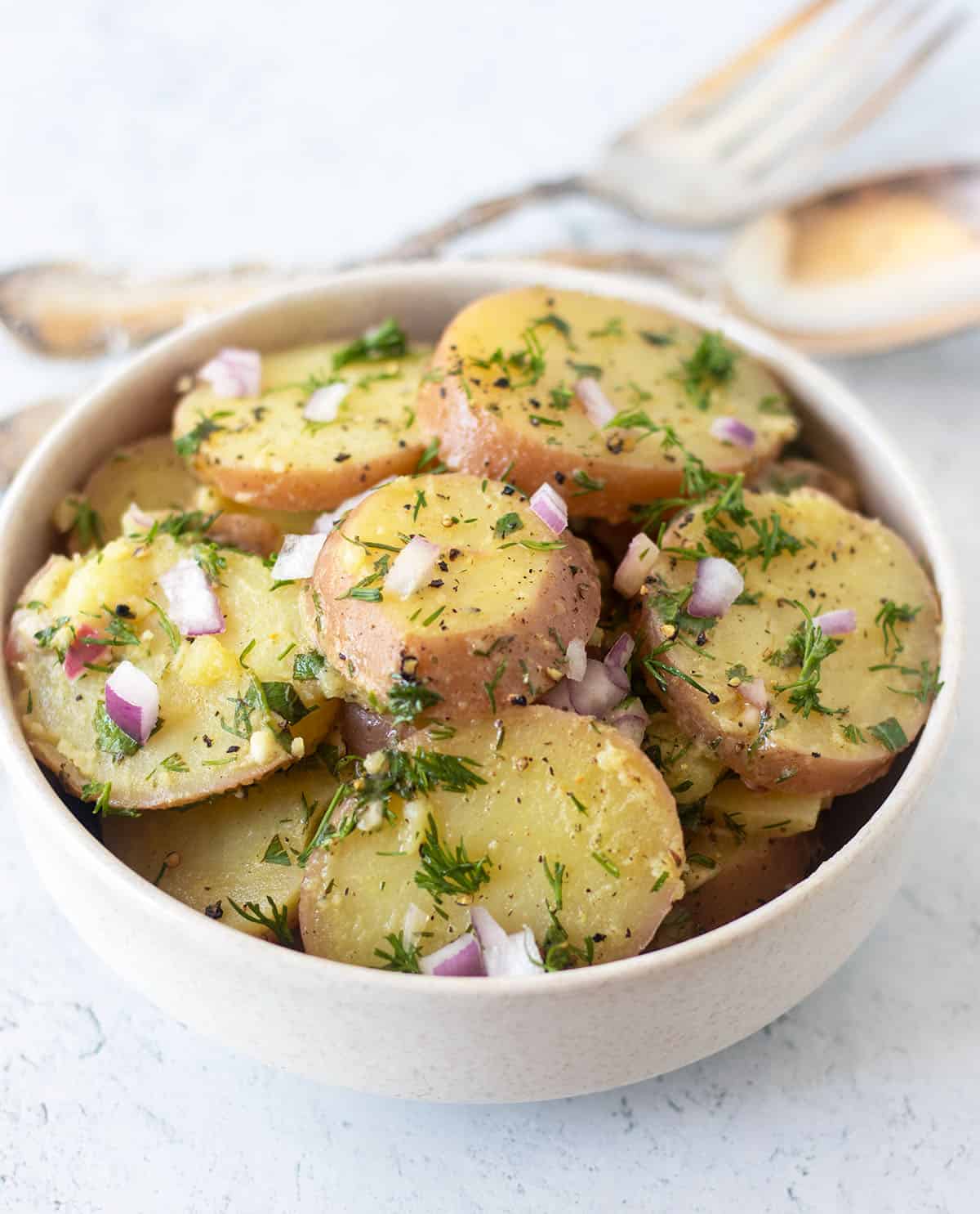 Vegan Dill potato salad in a white bowl with serving fork & spoon in background.