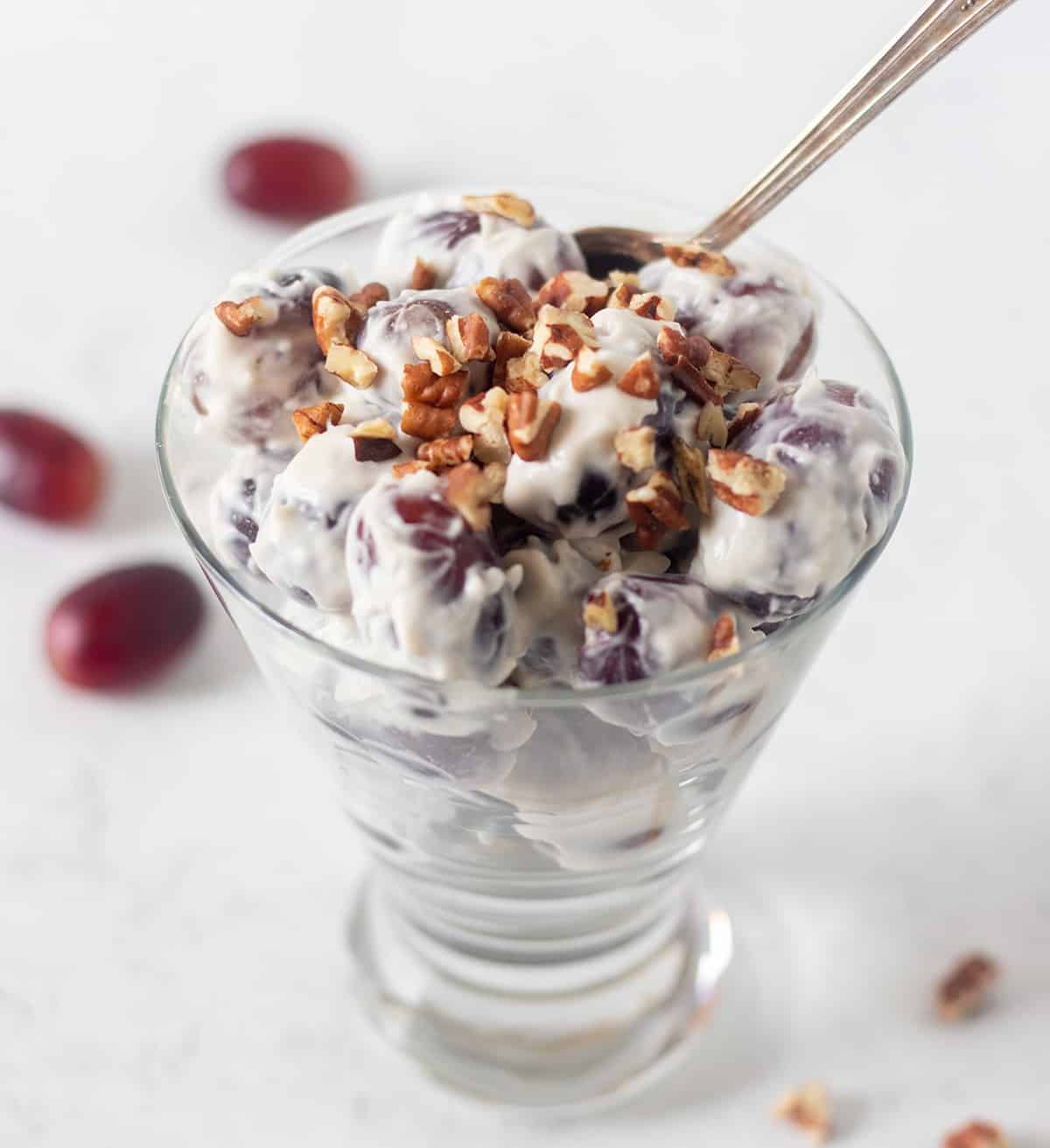 Grape fruit salad in a clear dessert dish with a silver spoon topped with chopped pecans. Red grapes in the background.