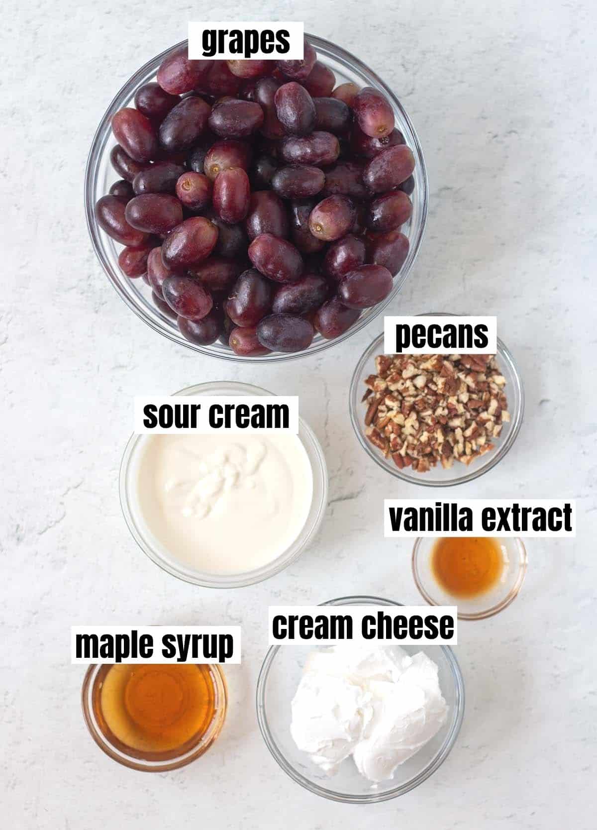 grape salad ingredients including red grapes, sour cream, chopped pecans, vanilla extract, maple syrup, & cream cheese.
