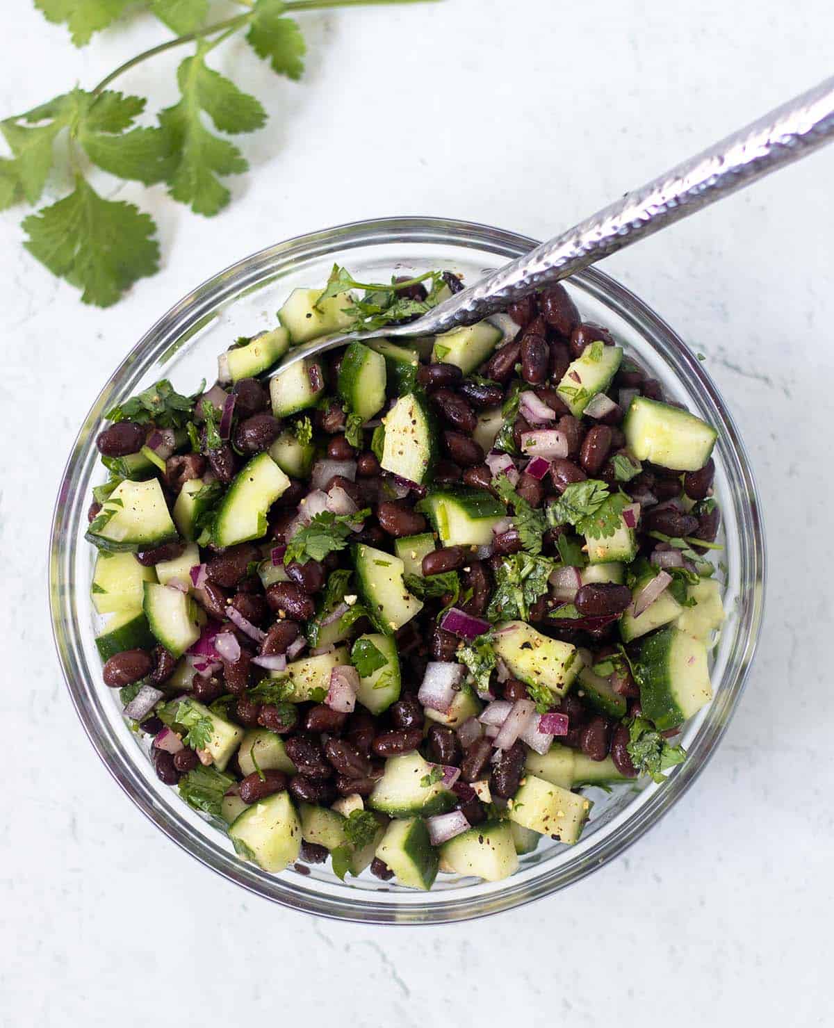 cucumber black bean salad in a clear glass bowl with a silver spoon for serving. Cilantro leaves in the background.