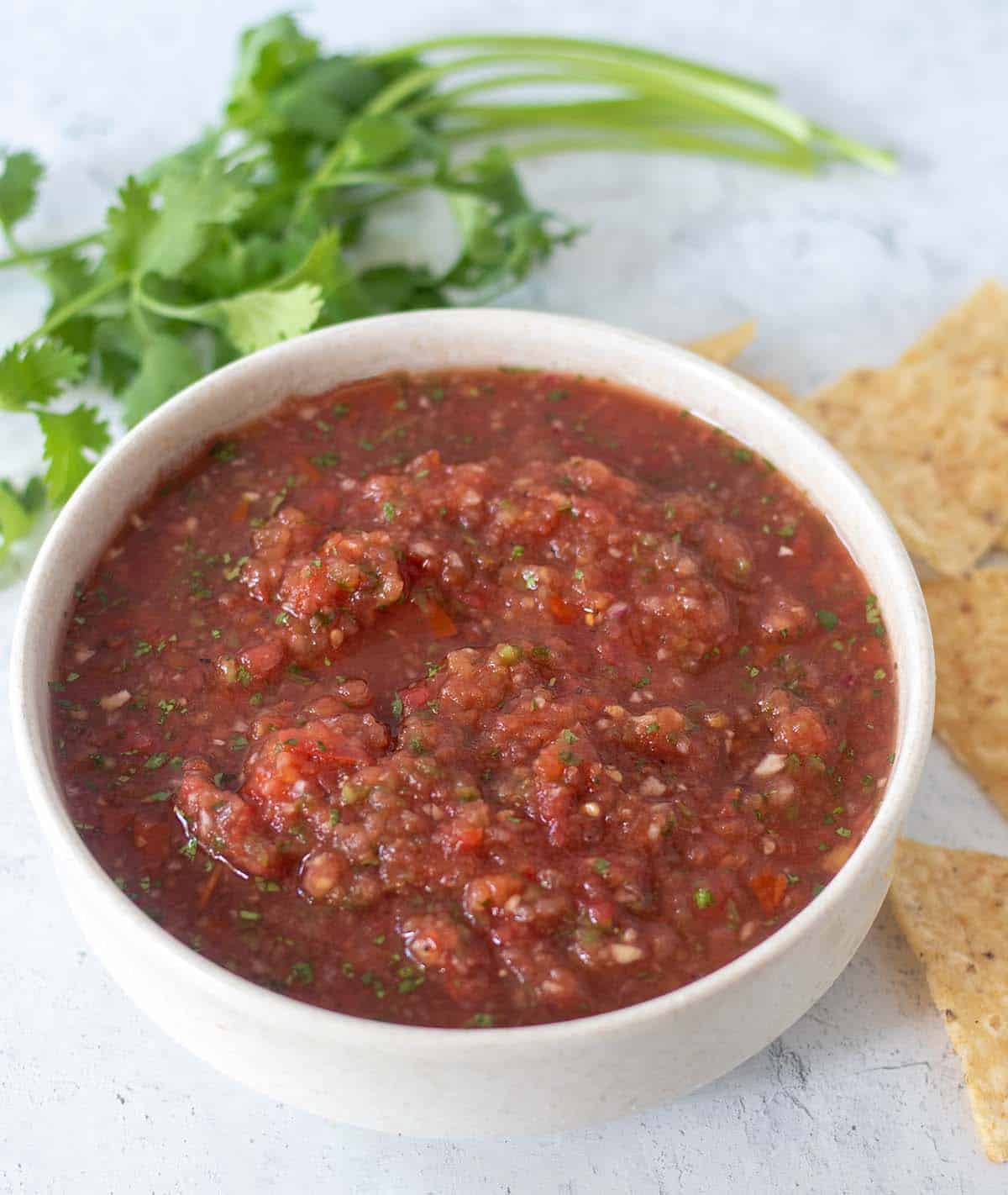 Homemade salsa in a white bowl with cilantro and tortilla chips in the background.