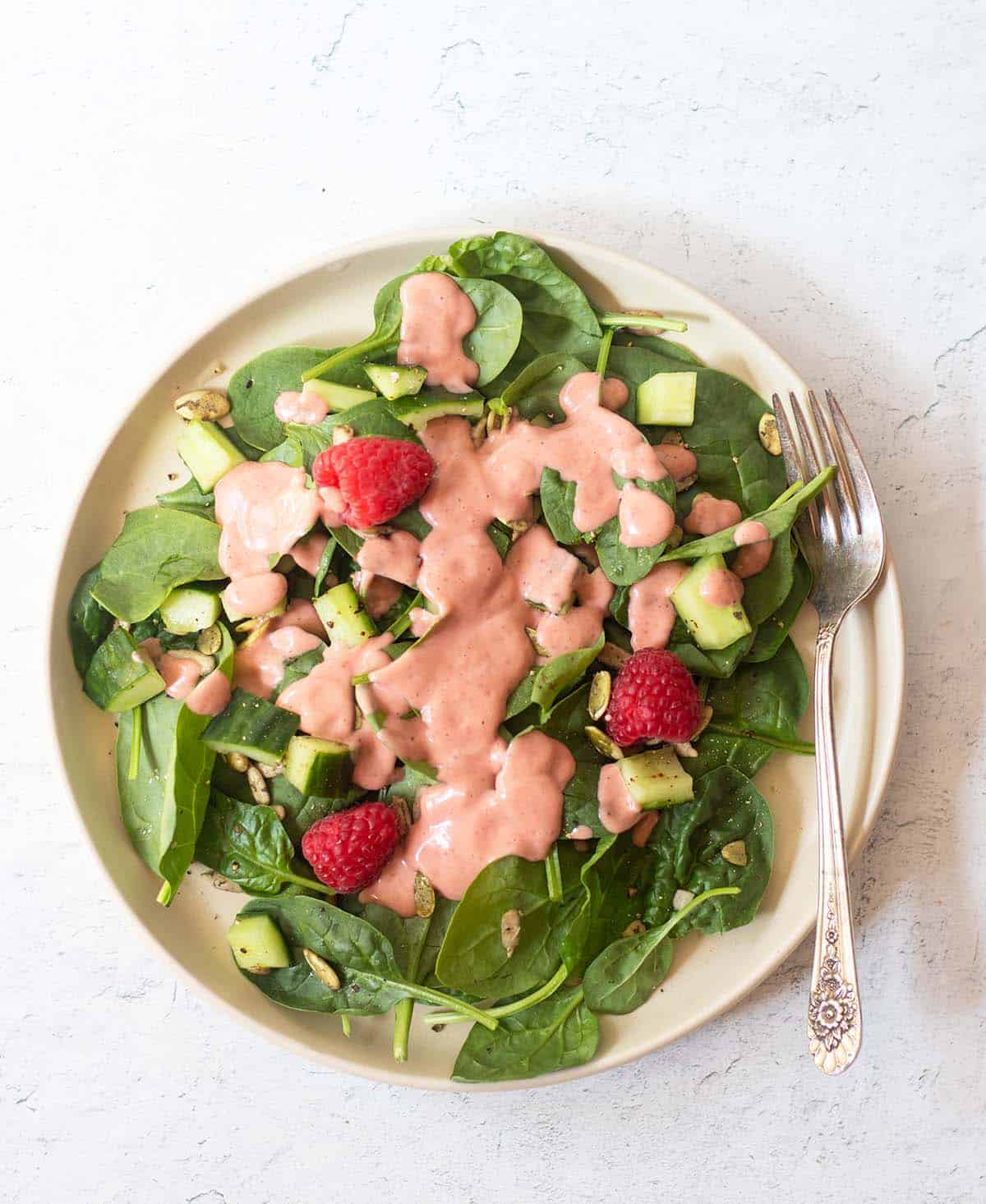 Raspberry Vinaigrette drizzled on top of a spinach salad with cucumbers and fresh raspberries. It's served on a beige salad plate with a silver fork.