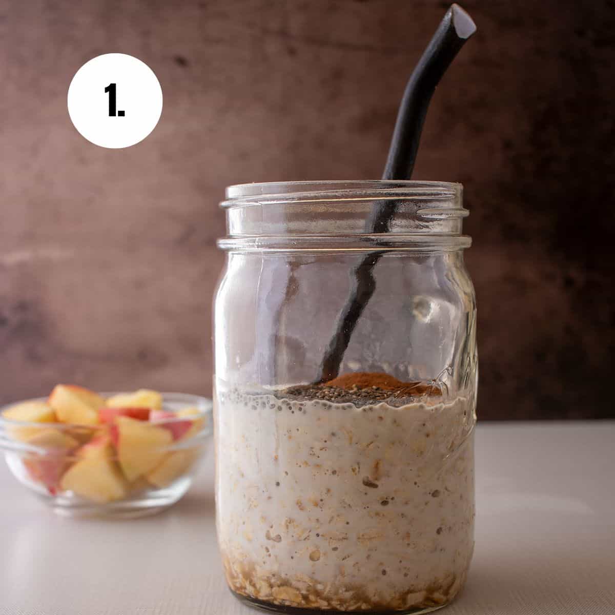 apple cinnamon overnight oats ingredients in a glass jar with a spoon. Apples in a glass bowl in the background.