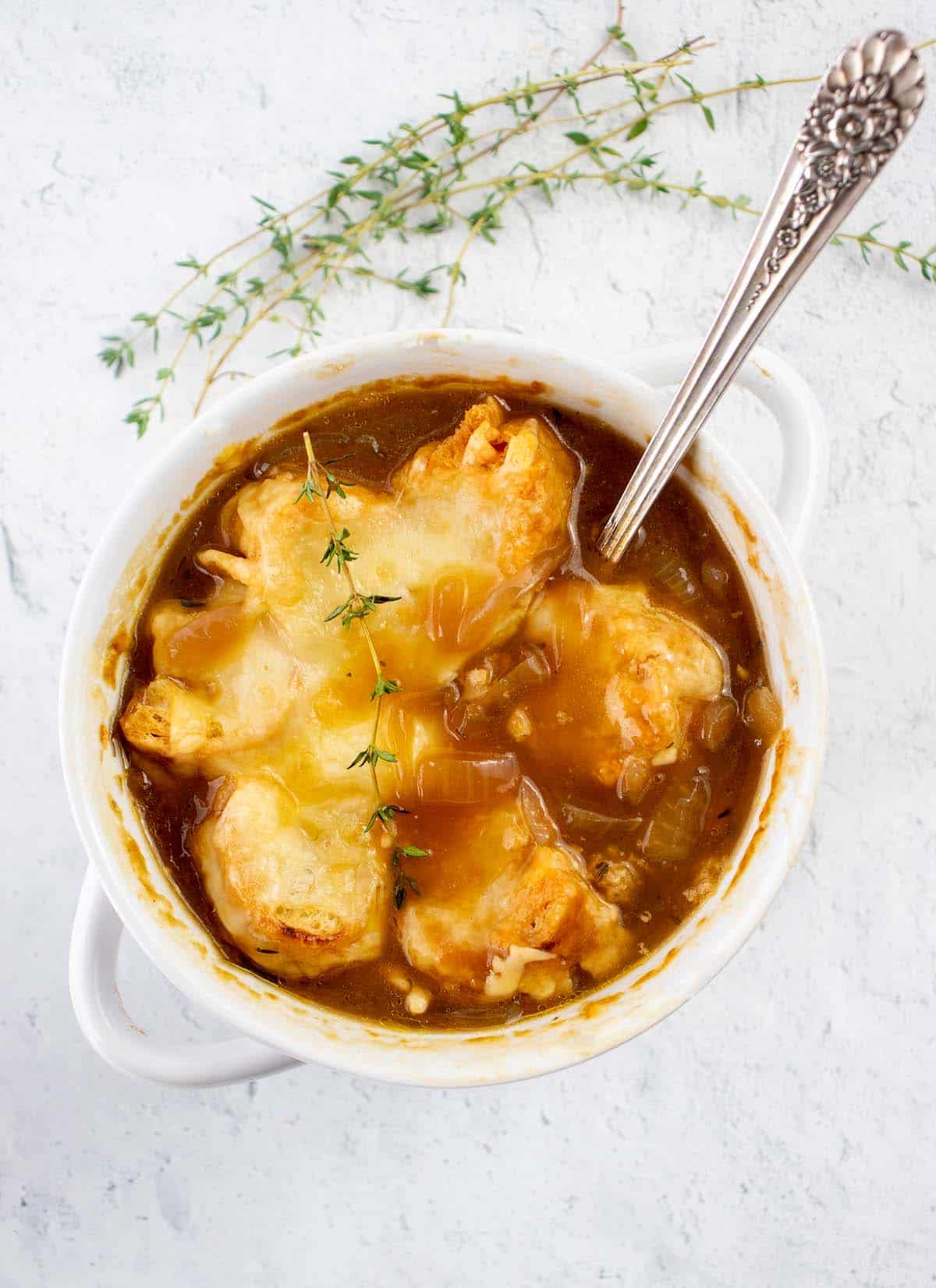 Vegetarian french onion soup in a white bowl with cheesy crouton topping and a silver spoon. Fresh thyme on top for garnish and beside the bowl.