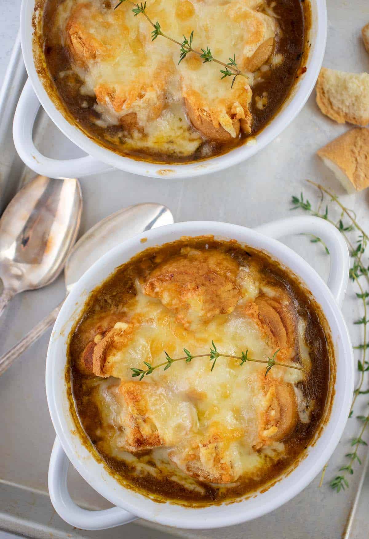 Vegetarian french onion soup in white bowls topped with melted cheese, and croutons on a silver tray with spoons, croutons and fresh thyme.