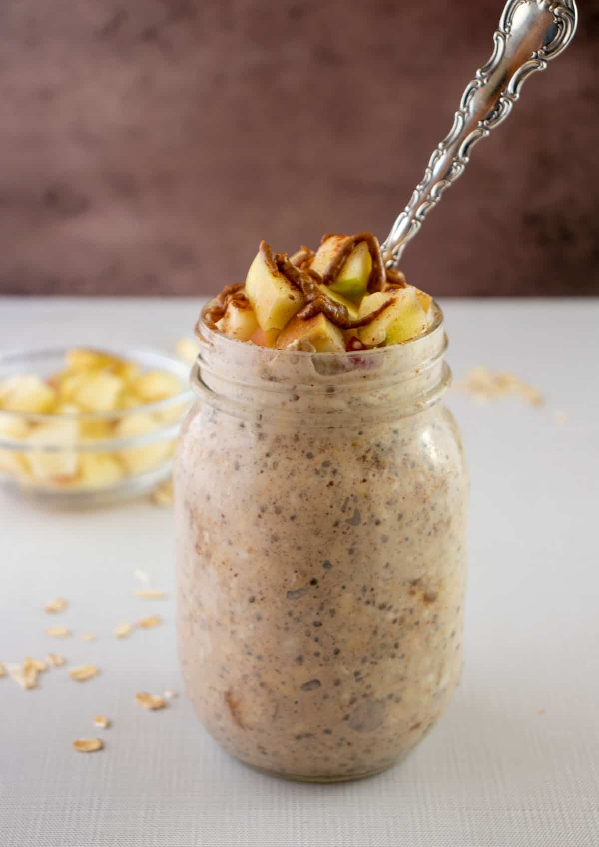 apple cinnamon overnight oats in a glass mason jar with a silver spoon topped with apples, cinnamon and almond butter. A bowl of chopped apples in the background. And oats laying next to the jar.