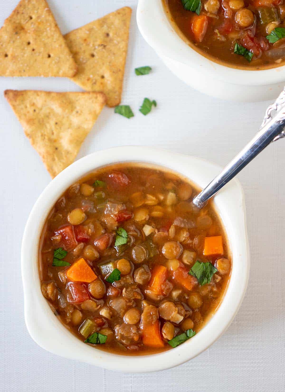 slow cooker lentil soup in a white soup crock with a silver spoon. Crackers next the bowl with chopped parsley.