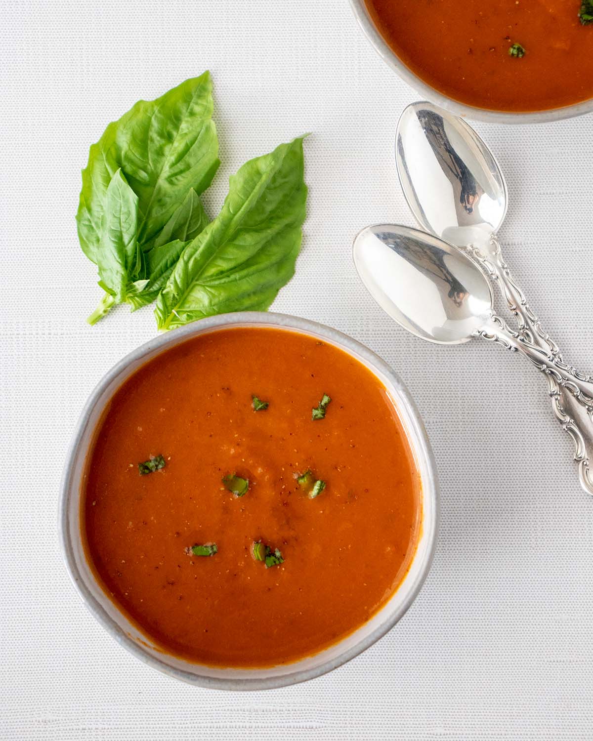 Vegan tomato soup in a grey bowl topped with fresh basil and cracked black pepper. Fresh basil leaves and two silver spoons beside the bowl.