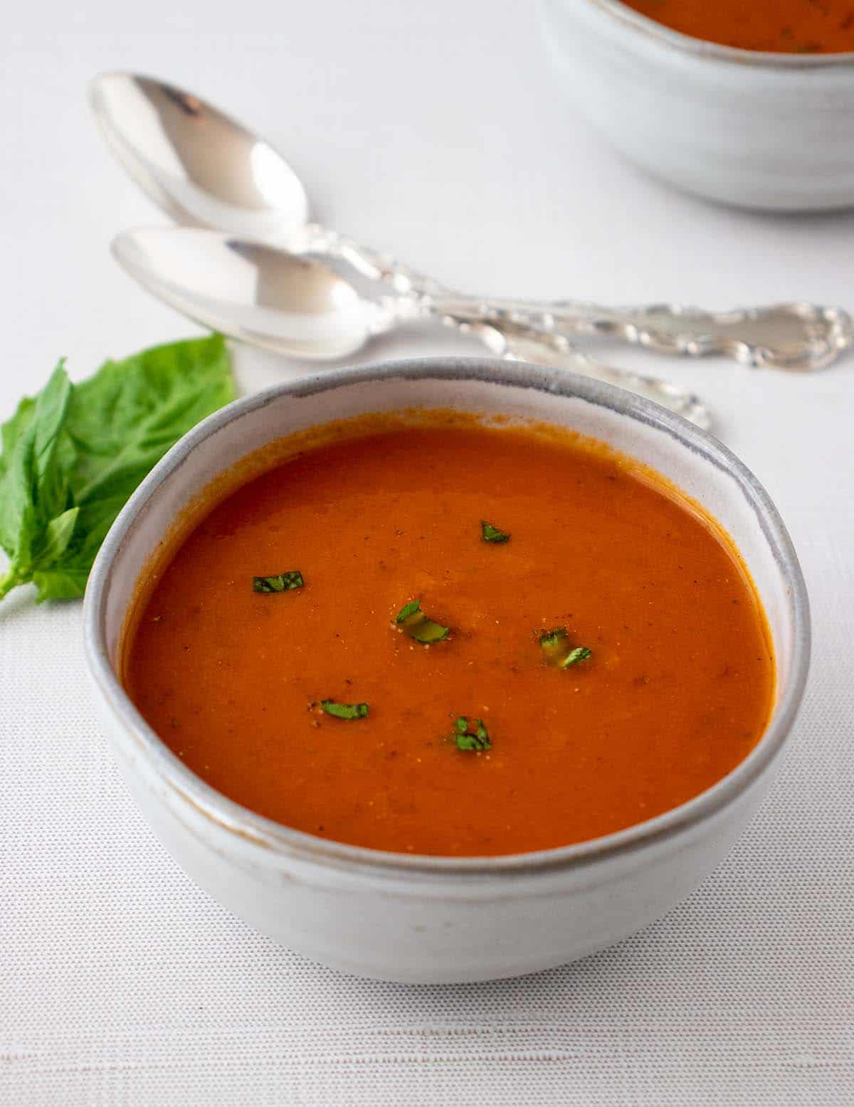 vegan tomato soup in a grey bowl garnished with chopped fresh basil and black pepper. Fresh basil in the background along with silver spoons.