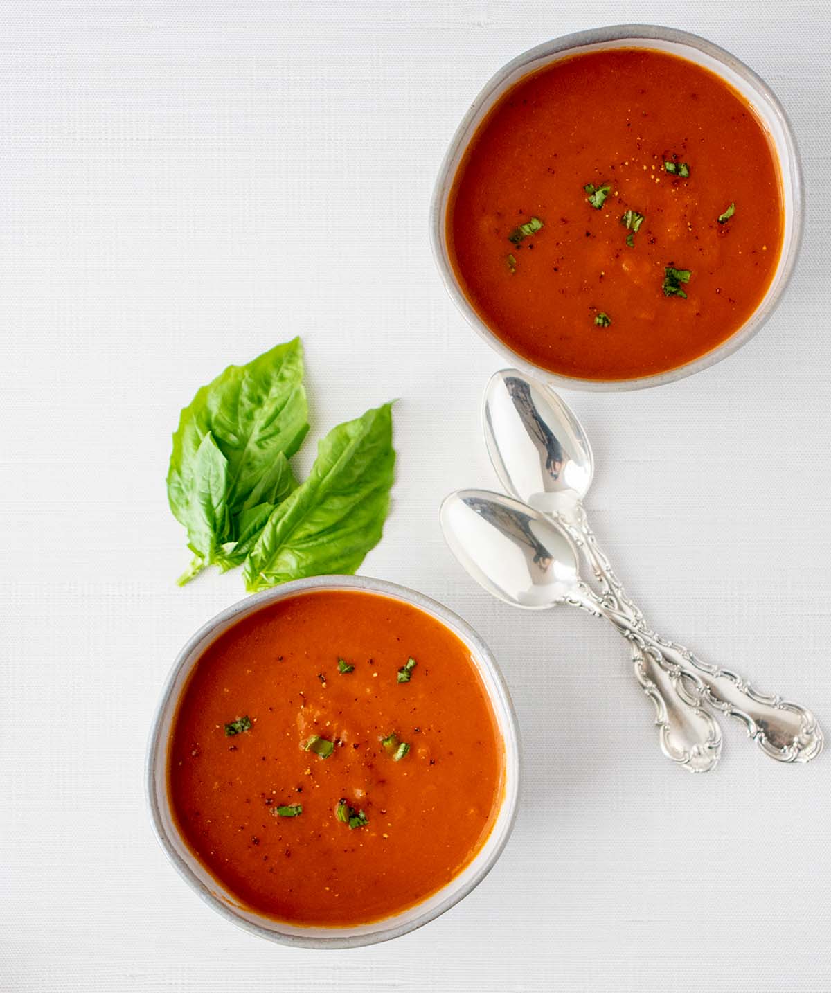 Two bowls of vegan tomato soup with silver spoons. Soup is garnished with fresh basil and black pepper. Fresh basil in the background.
