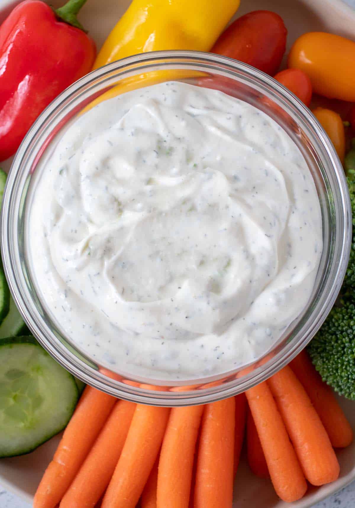 Homemade ranch dip in a clear glass bowl on a plate of veggies.