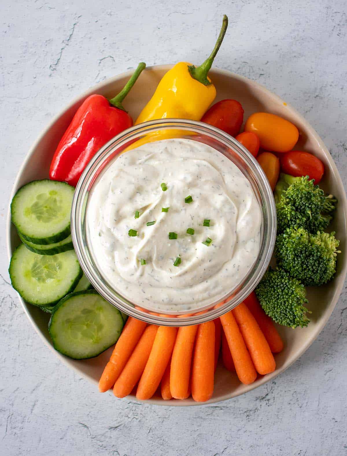 Homemade ranch dip in clear glass bowl topped with fresh chives surrounded by a plate of fresh cut veggies. Carrots, broccoli, cucumber, tomatoes and peppers.