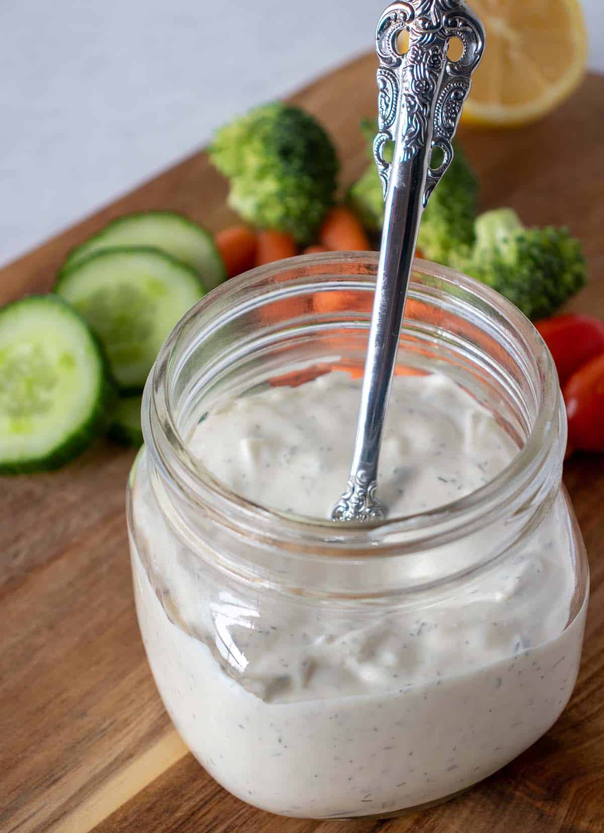 Homemade ranch dip in a clear glass jar with a silver spoon. Fresh veggies including cucumber slices, broccoli, carrots, tomatoes and a lemon slice in the background.
