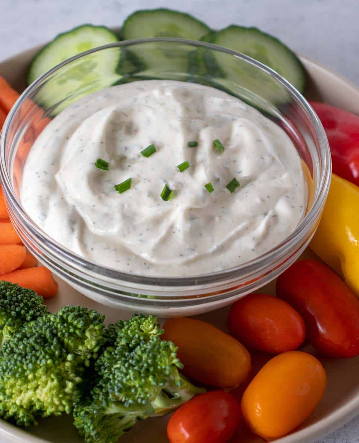 homemade ranch dip in a clear glass bowl garnished with fresh chopped chives. Fresh veggies surrounding the bowl which include tomatoes, broccoli, carrots, cucumbers and bell peppers.