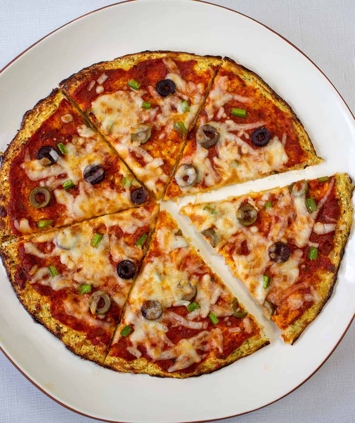 Cauliflower pizza crust that's been topped with tomato sauce, olives, bell peppers and cheese that's been baked and sliced. Pizza is on a white serving plate with brown trim.