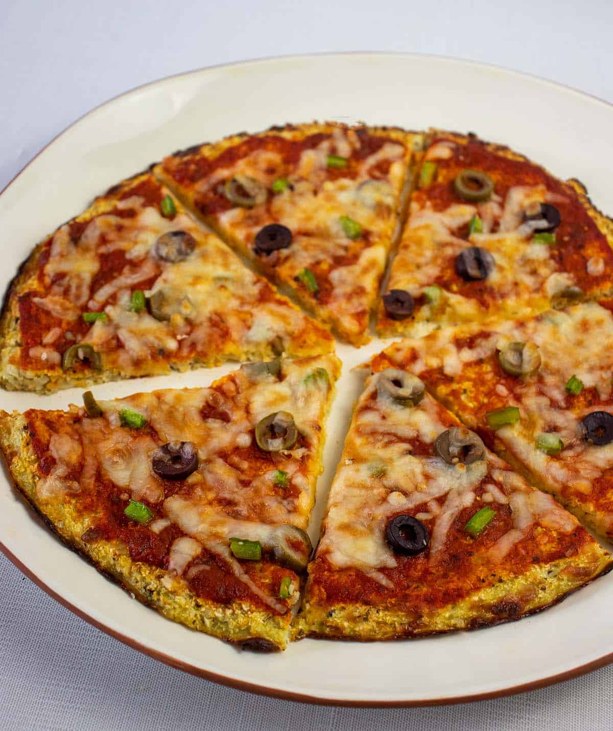 Cauliflower pizza crust topped with tomato sauce, olives, bell peppers and mozzarella cheese on a white plate with brown trim. Pizza is sliced in diagonal slices.