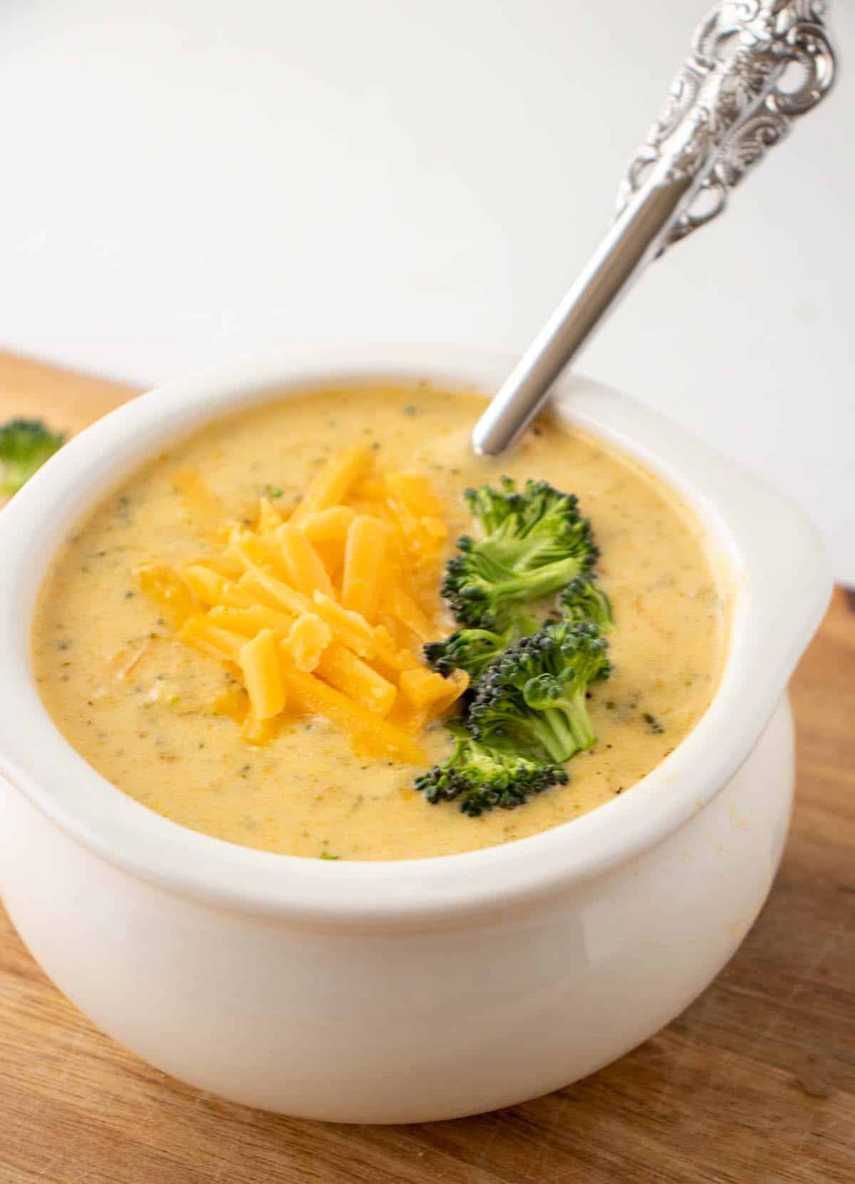 broccoli cheddar soup in a white bowl topped with shredded cheddar cheese and broccoli florets sitting on a brown cutting board. A silver spoon in the bowl for serving.