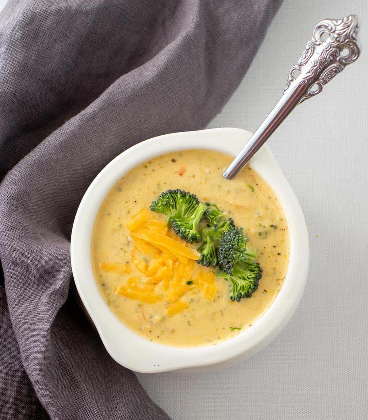 Instant pot broccoli cheddar soup in a white bowl topped with shredded cheddar cheese and broccoli florets. A silver spoon in bowl and a grey napkin next to the bowl.