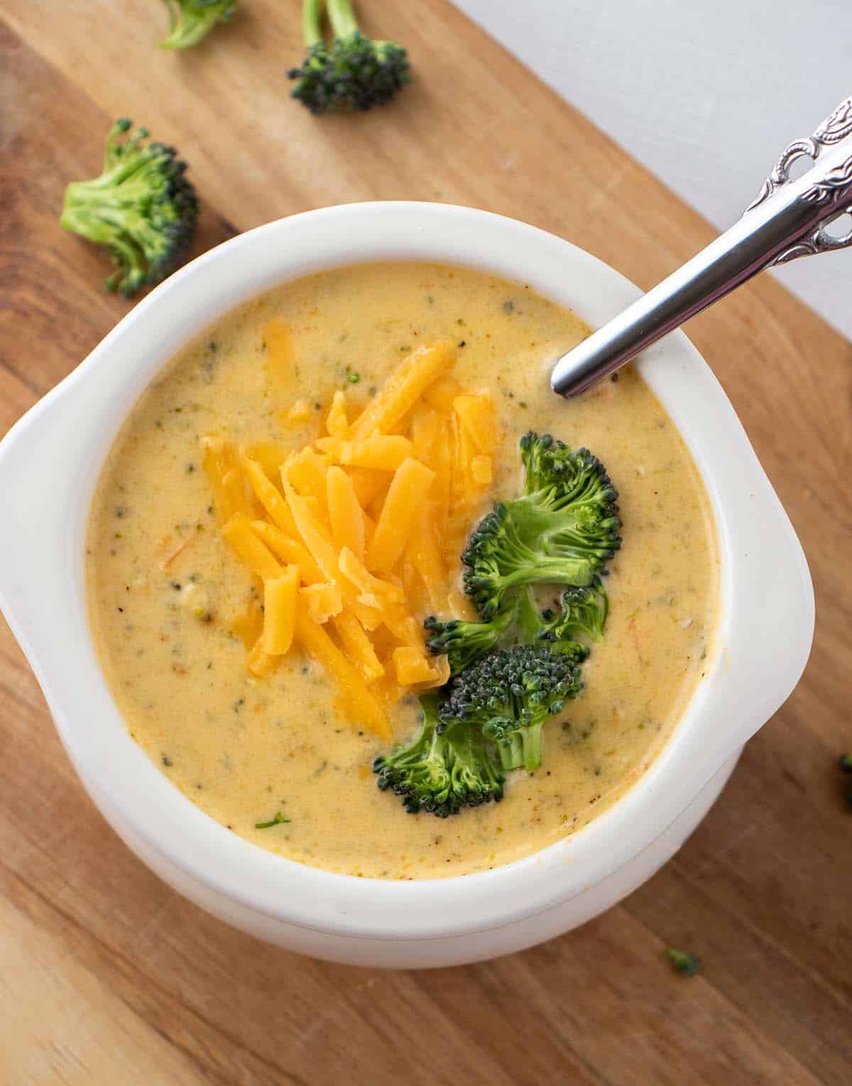 Instant pot broccoli cheddar soup in a white bowl topped with shredded cheddar cheese and broccoli florets. A silver spoon in the bowl and broccoli florets next to the bowl.