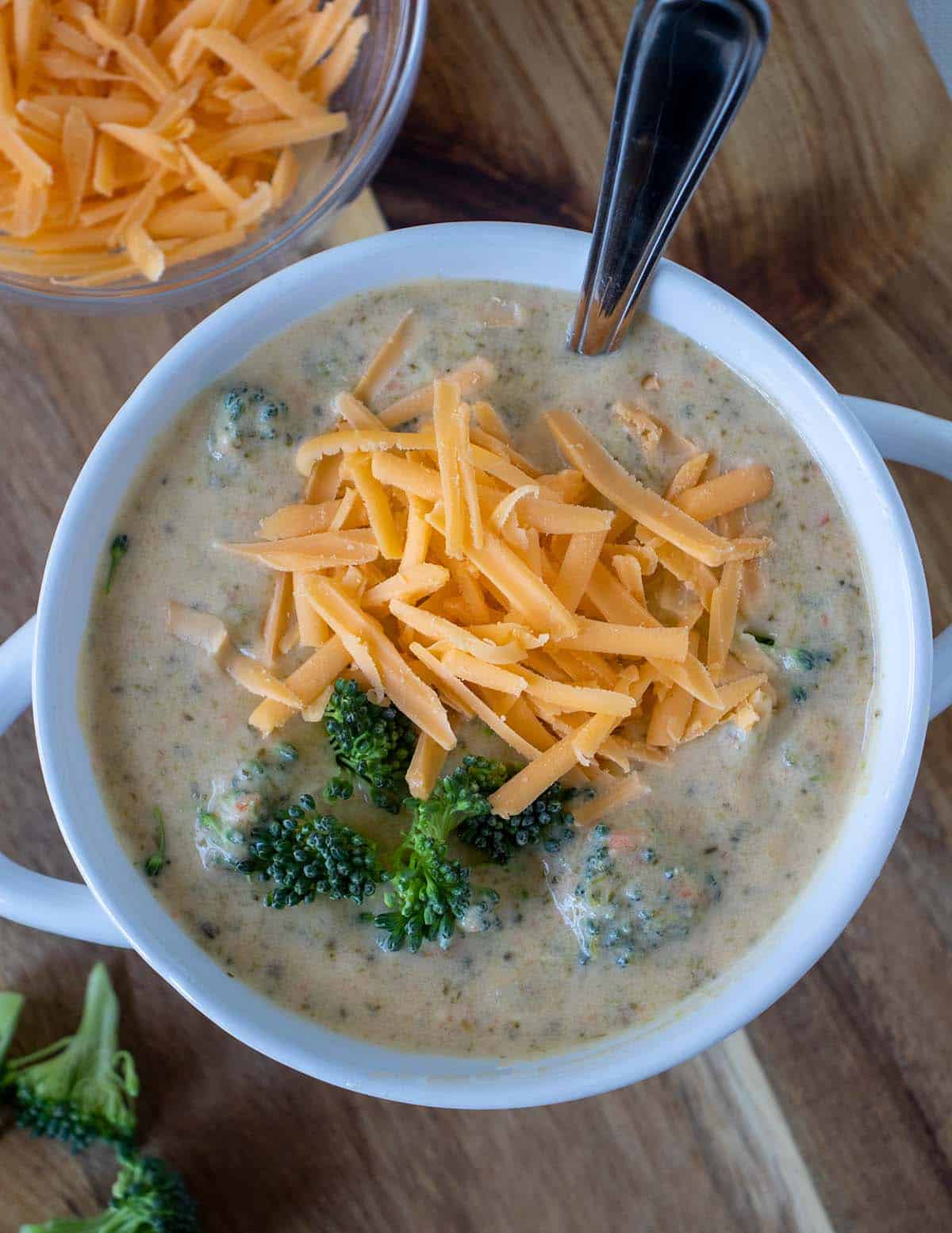 Easy broccoli cheddar soup in a white crock with a silver spoon topped with shredded cheddar cheese and fresh broccoli florets. a bowl of shredded cheddar cheese beside the soup.