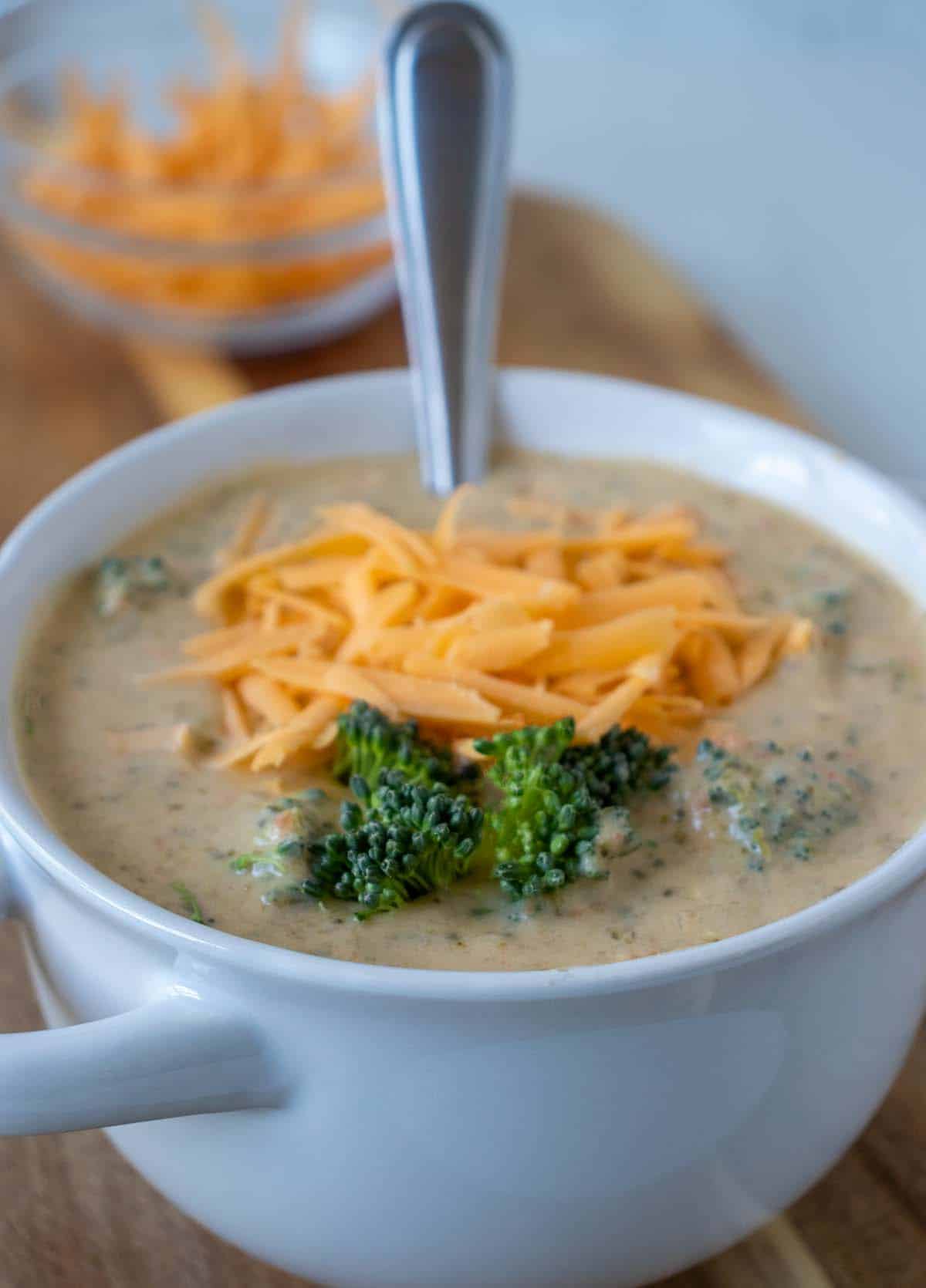 Broccoli cheddar soup in a white crock on a brown cutting board that's topped with shredded cheddar cheese and broccoli florets.